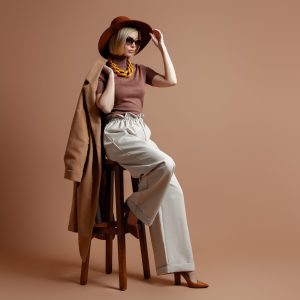 Fashionable young woman in elegant hat sitting on a chair against brown background