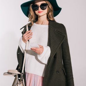 beautiful teenage girl in black hat and sunglasses using smartphone isolated on grey